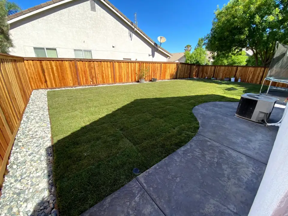 Curved green backyard lawn beside a concrete patio, pale rock border, and wood fence