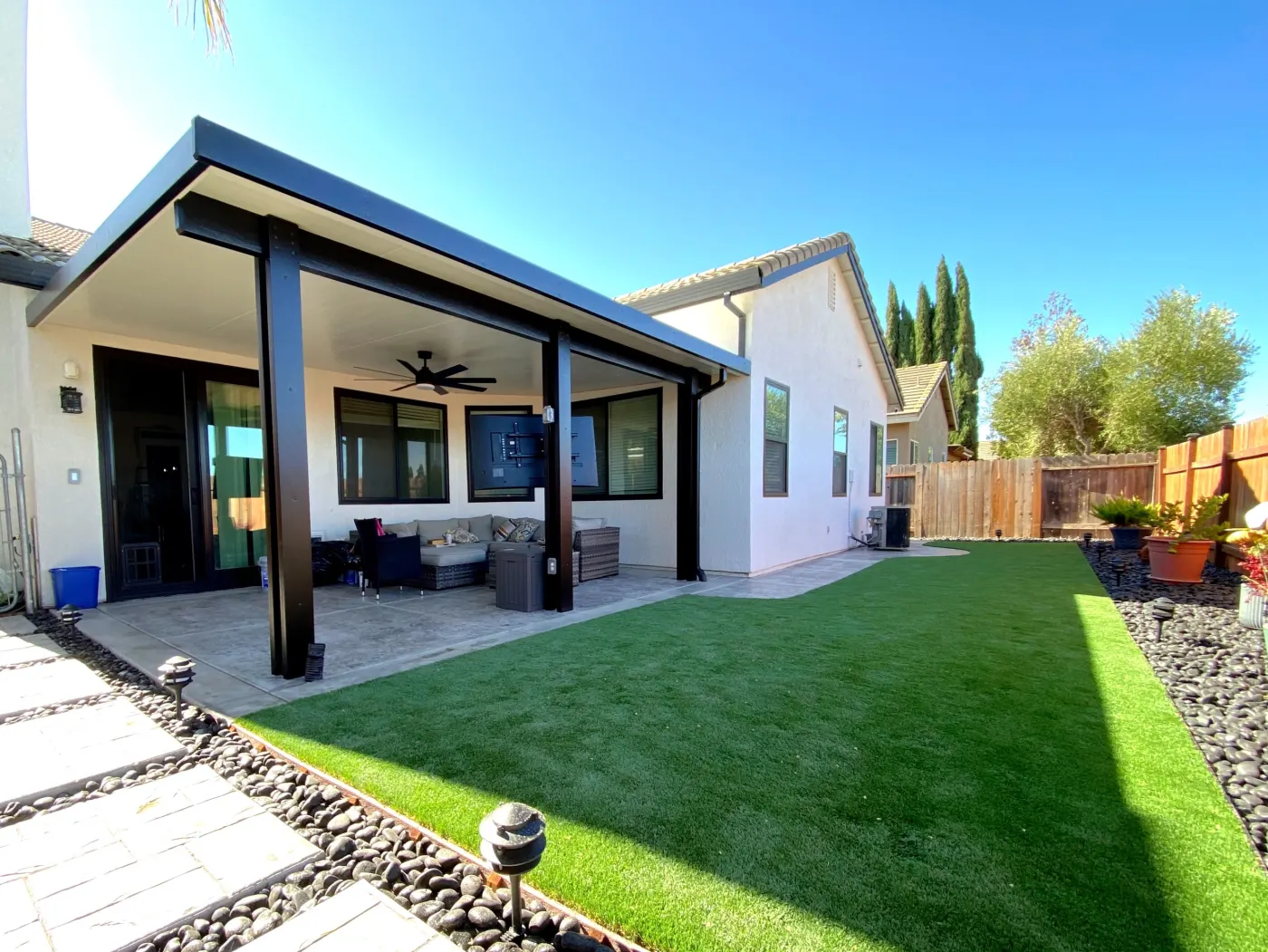 Covered backyard patio with black posts, ceiling fans, green lawn, and a paved walkway