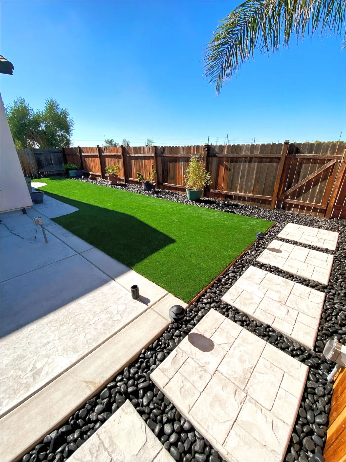 Backyard with bright green turf, concrete patio, square pavers, dark rock borders, and wood fencing