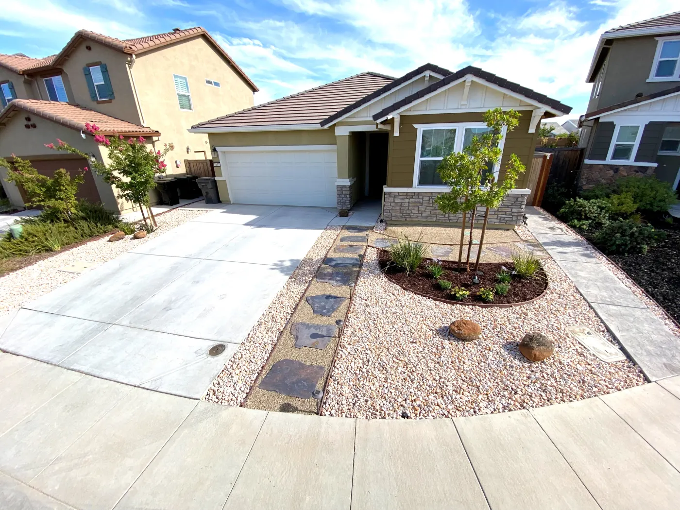 Front yard with a wide concrete driveway, stone ground cover, stepping path, and young trees