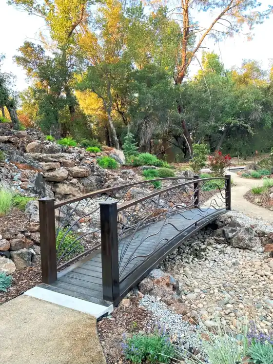 Small garden bridge with black metal railings crossing a dry rock creek beside a path