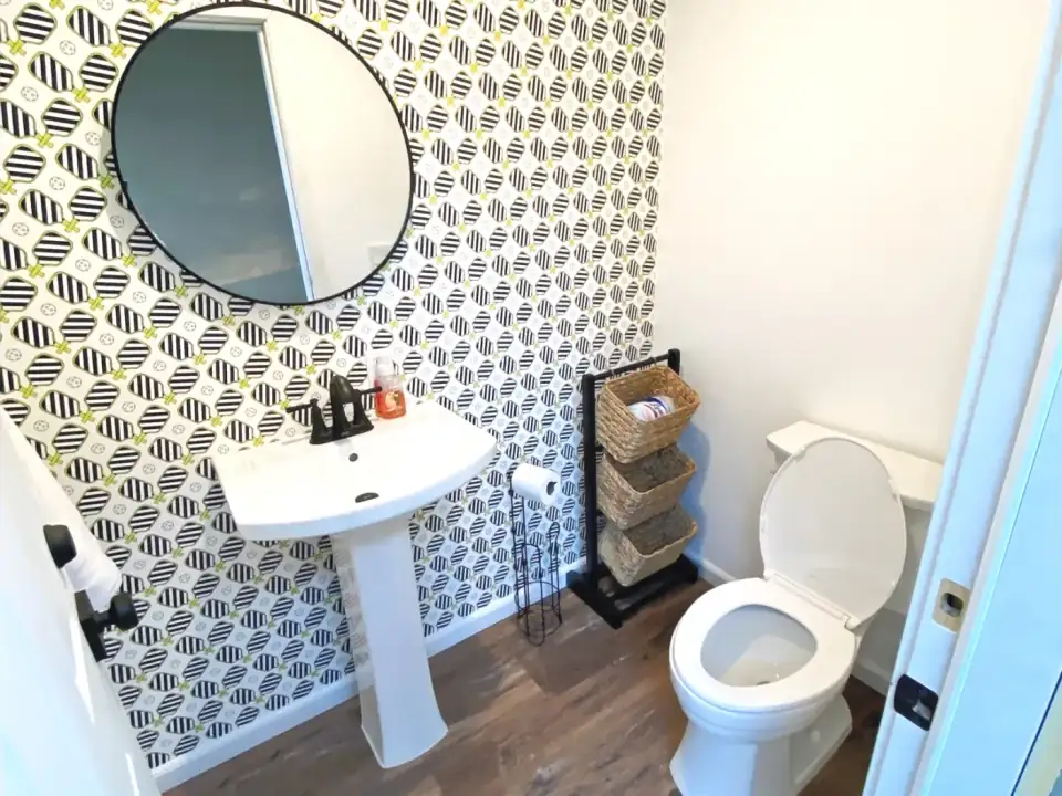 Powder room with black-and-white patterned wallpaper, round mirror, pedestal sink, and white toilet